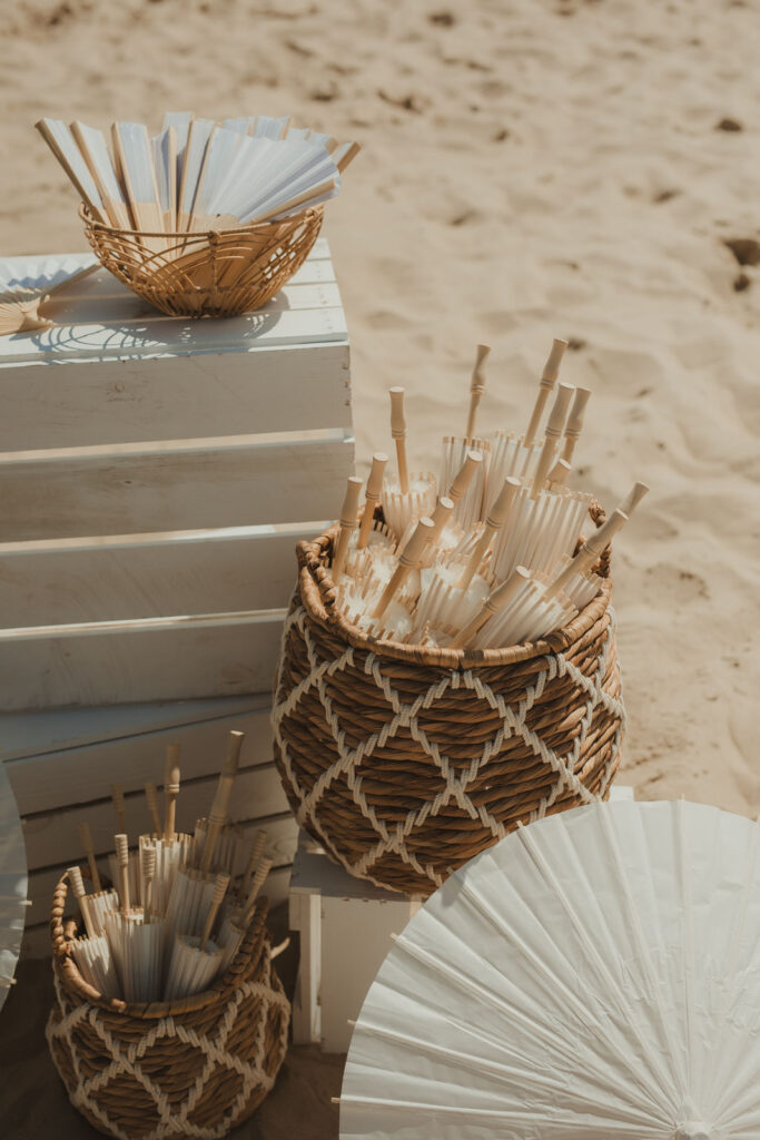 Geflochtene Körbe, gefüllt mit weißen Papierfächern, ruhen auf Holzkisten an einem sandigen Ostseestrand.