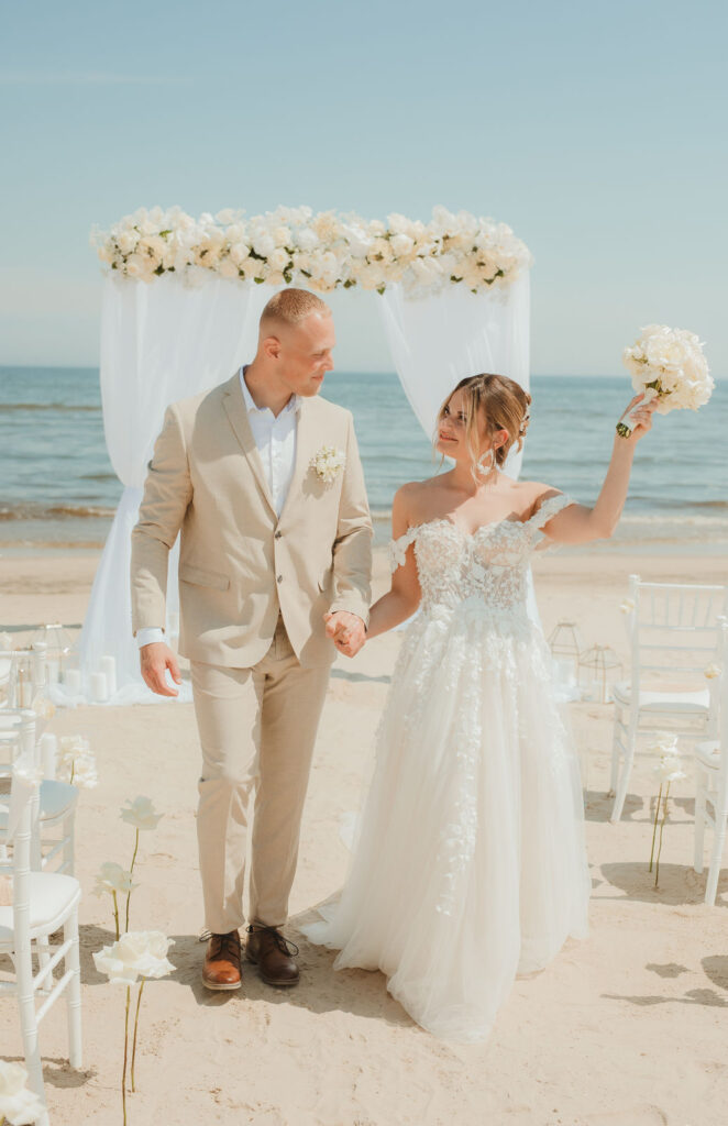 Braut und Bräutigam halten sich am Strand von Rügen an den Händen, mit einem Blumenbogen und Stühlen; die Braut hebt ihren Brautstrauß unter dem sonnigen Himmel von Mecklenburg-Vorpommern.