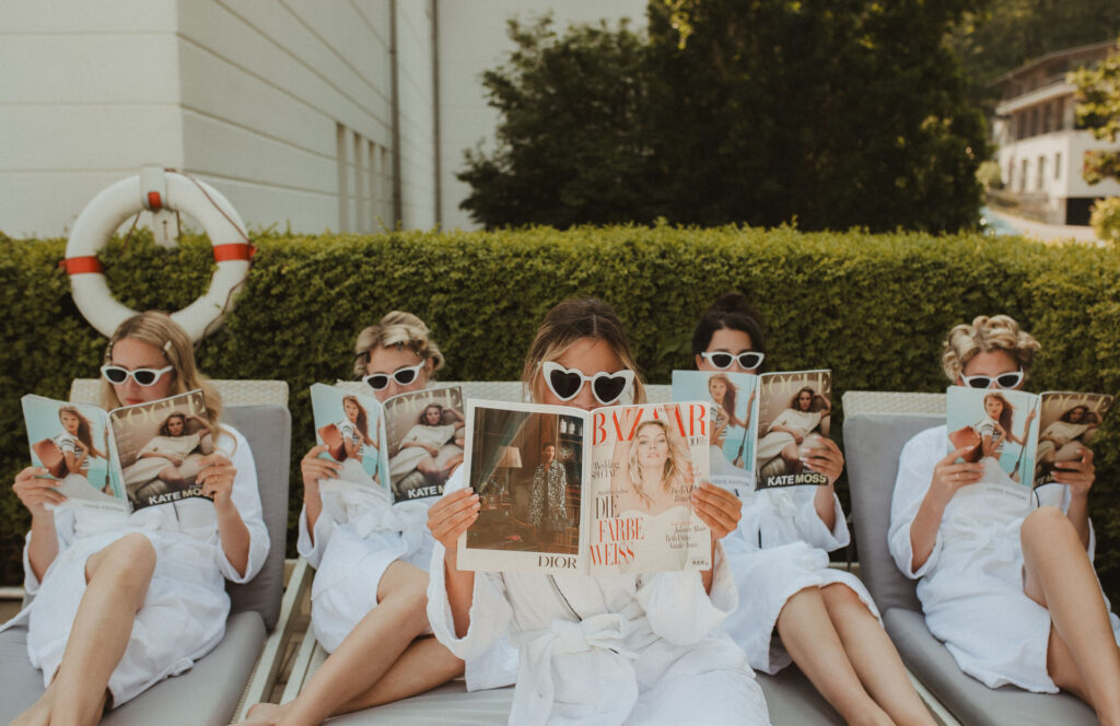 Fünf Frauen in weißen Bademänteln und mit Herz-Sonnenbrillen lesen Zeitschriften, während sie an einem Pool liegen und einen entspannten Tag in Rostock genießen.