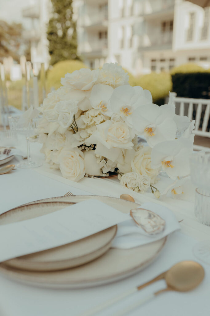 Elegant gedeckter Tisch mit Goldbesteck und einem Tafelaufsatz aus weißen Rosen und Orchideen, perfekt für eine anspruchsvolle Feier an der Ostsee oder unter Anleitung eines Hochzeitsplaners in Mecklenburg-Vorpommern - Heiraten an der Ostsee