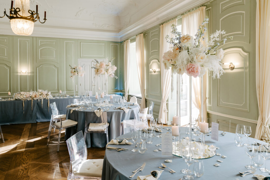 Elegante Hochzeitslocation in Mecklenburg-Vorpommern mit pastellfarbenem Dekor, floralen Tafelaufsätzen auf runden Tischen und großen Fenstern mit Vorhängen - perfekt für eine stilvolle Hochzeit an der Ostsee.