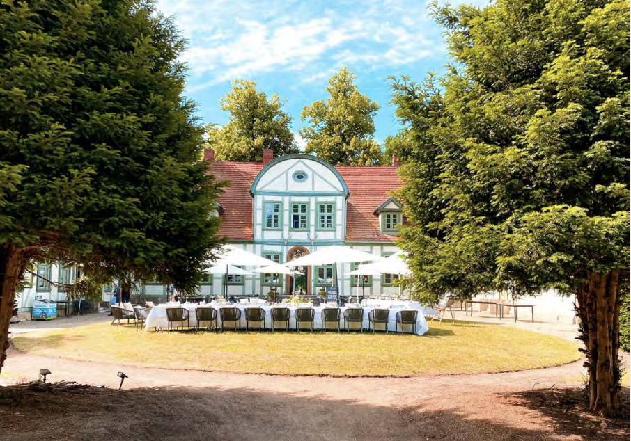 Vor einem großen, historischen Haus, umrahmt von hohen Bäumen, steht ein langer Esstisch mit weißen Sonnenschirmen - eine idyllische Hochzeitslocation in Mecklenburg-Vorpommern nahe der Ostsee.