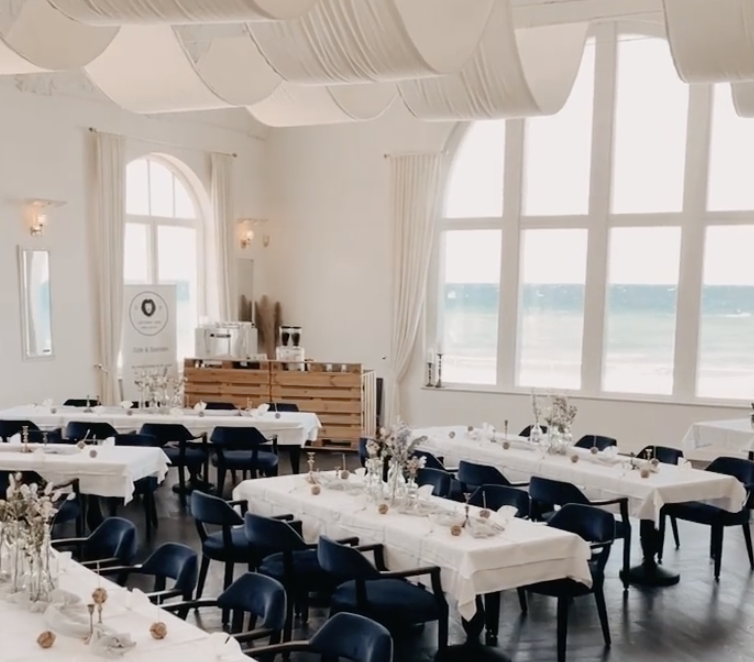 Eleganter Speisesaal mit weißen Tischtüchern, marineblauen Stühlen, großen Bogenfenstern und Meerblick - eine ideale Hochzeitslocation an der Ostsee in Mecklenburg-Vorpommern.