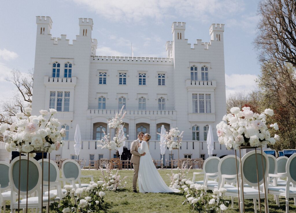 Braut und Bräutigam umarmen sich im Freien vor einem weißen Schloss mit floraler Hochzeitsdekoration, der perfekten Hochzeitslocation in Mecklenburg-Vorpommern nahe der Ostsee.