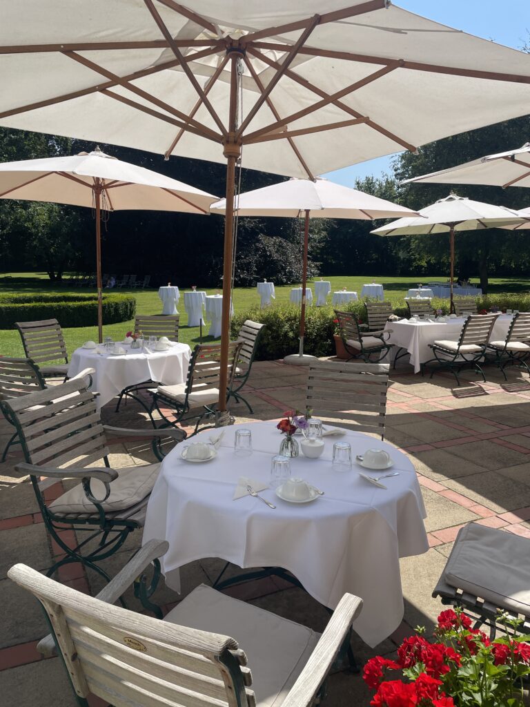 Eine Außenterrasse mit runden Tischen zum Essen, Holzstühlen und großen weißen Sonnenschirmen an einem sonnigen Tag - eine einladende Hochzeitslocation nahe der Ostsee im schönen Mecklenburg-Vorpommern.