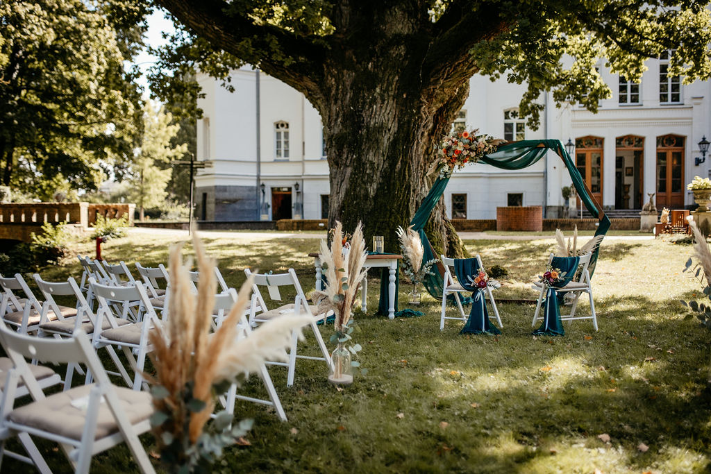 Hochzeit im Freien mit weißen Stühlen, Pampasgras und einem dekorierten sechseckigen Bogen unter einem großen Baum - eine idyllische Hochzeitslocation an der Ostsee in Mecklenburg-Vorpommern.