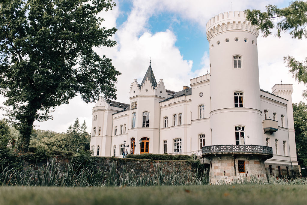 Ein weißes Schloss mit hohen Türmen und gewölbten Fenstern, umgeben von Bäumen unter einem teilweise bewölkten Himmel, bietet eine romantische Hochzeitslocation in Mecklenburg-Vorpommern nahe der Ostsee.