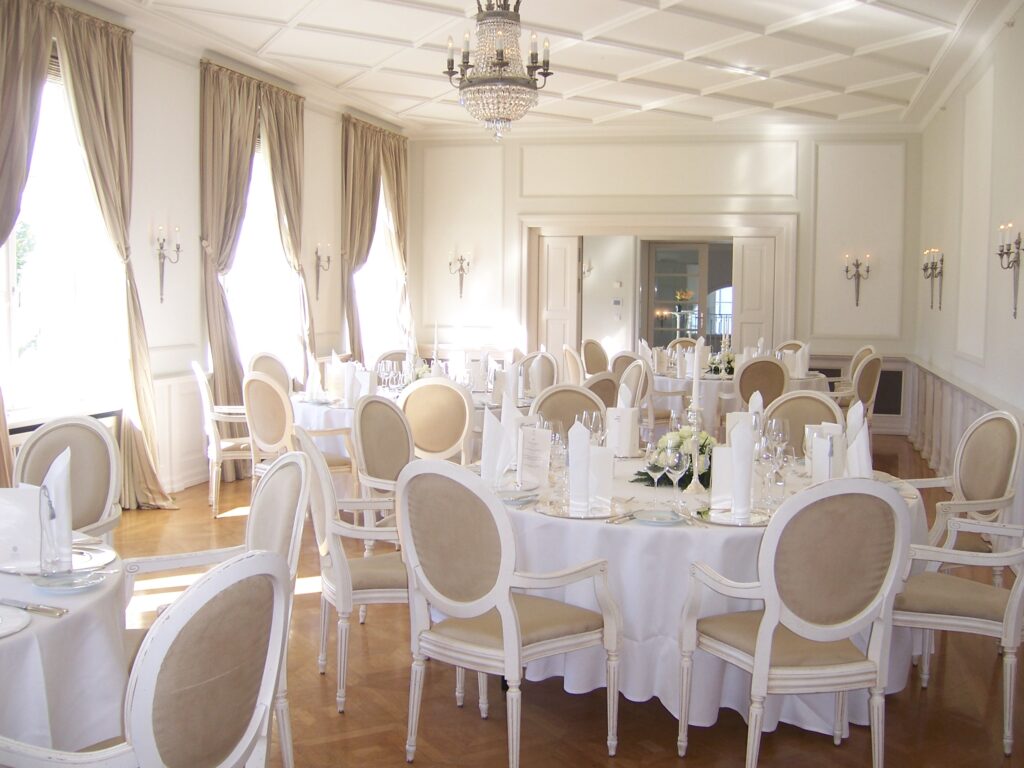 Eleganter Festsaal mit runden Tischen, weißen Stühlen, Kronleuchtern und großen Fenstern mit beigen Vorhängen - eine ideale Hochzeitslocation nahe der Ostsee in Mecklenburg-Vorpommern.