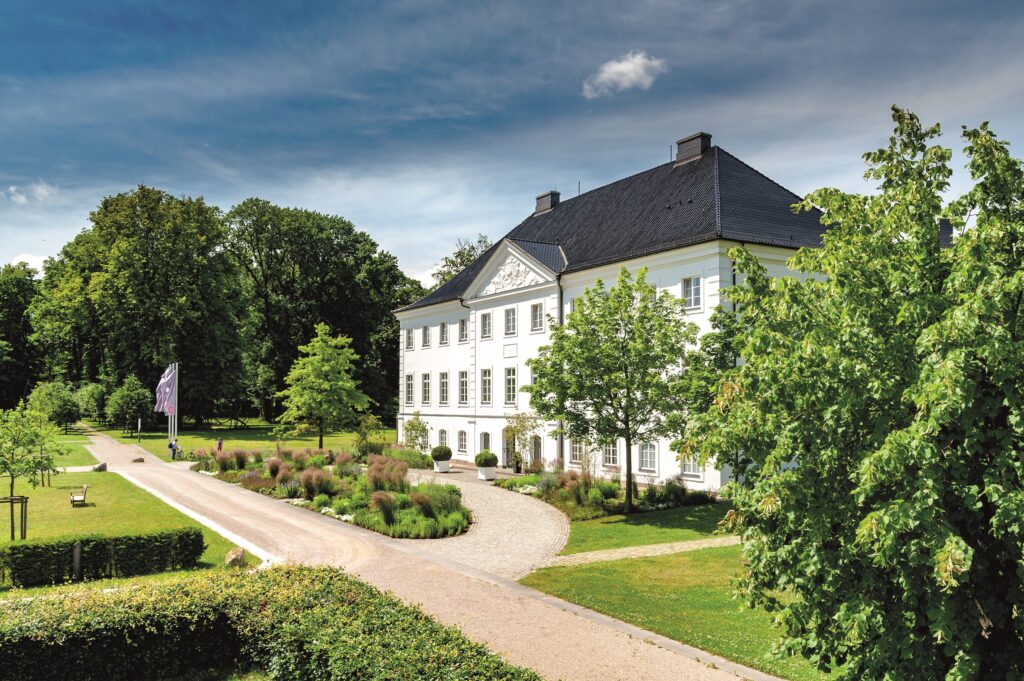 Charmantes weißes historisches Gebäude in Mecklenburg-Vorpommern, umgeben von grünen Bäumen und Rasen unter einem teilweise bewölkten blauen Himmel - eine ideale Hochzeitslocation in der Nähe der Ostsee.