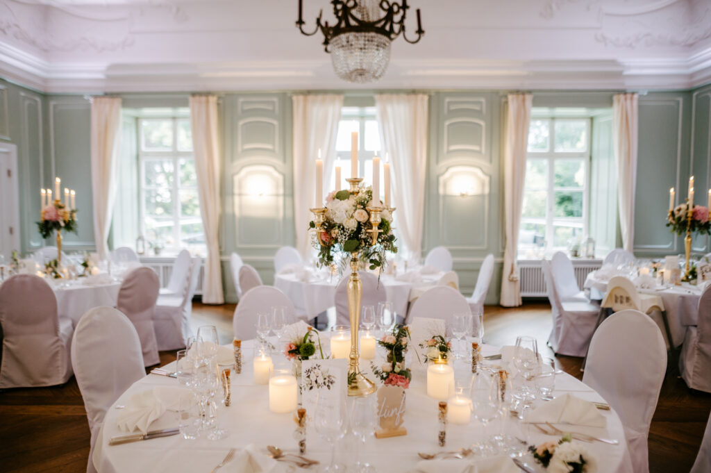 Elegante Hochzeitslocation in der Nähe der Ostsee mit rundem Tisch, Blumenschmuck, Kerzen und großen Fenstern im charmanten Mecklenburg-Vorpommern.