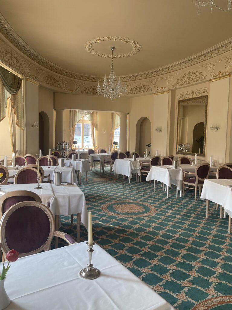 Eleganter Speisesaal mit runden Tischen, Kronleuchtern, verzierter Decke und großen Fenstern, die viel Licht hereinlassen - eine ideale Hochzeitslocation in Mecklenburg-Vorpommern, perfekt für unvergessliche Feiern an der Ostsee.