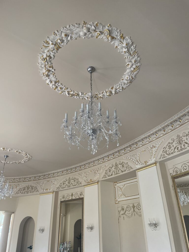 In einer luxuriösen Hochzeitslocation in Mecklenburg-Vorpommern hängt ein eleganter Kristalllüster von einer kunstvoll mit Weiß und Gold verzierten Decke herab.