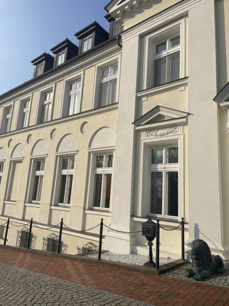 Elegantes cremefarbenes Gebäude mit großen Fenstern, Eisenzaun, gemauertem Gehweg und einer dekorativen Löwenstatue - ein idealer Hochzeitsort in Mecklenburg-Vorpommern nahe der Ostsee.