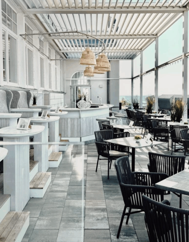 Ein modernes, luftiges Außencafé mit schwarzen Stühlen, weißen Tischen und Rattan-Hängeleuchten unter einer Pergola - eine ideale Hochzeitslocation an der Ostsee in Mecklenburg-Vorpommern.